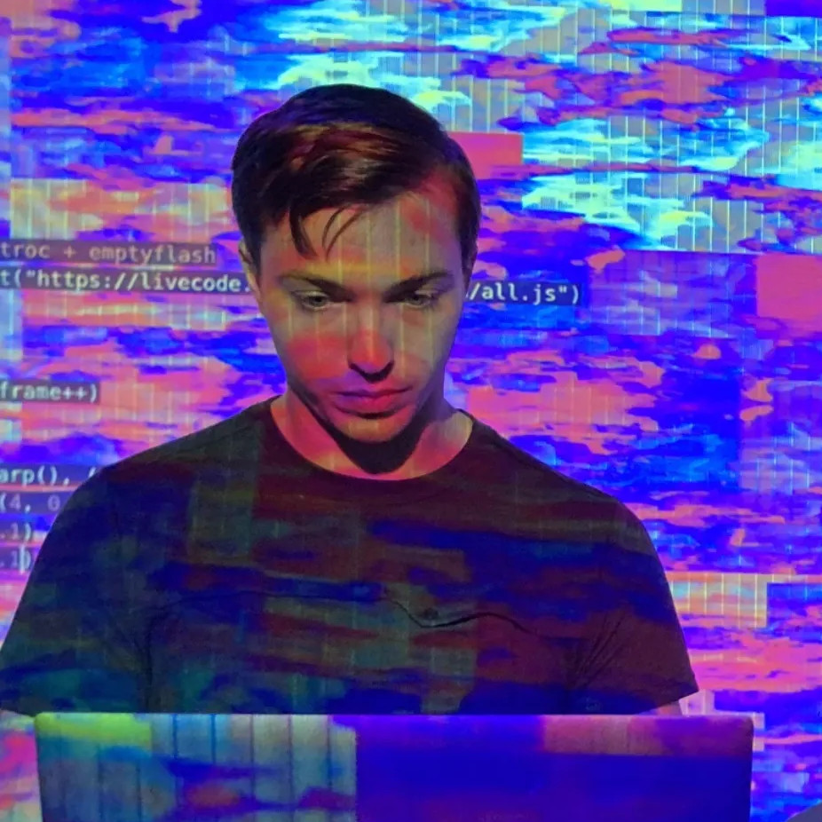 Close-up photo of emptyflash looking at his computer, with live coding projection on his face and background. 