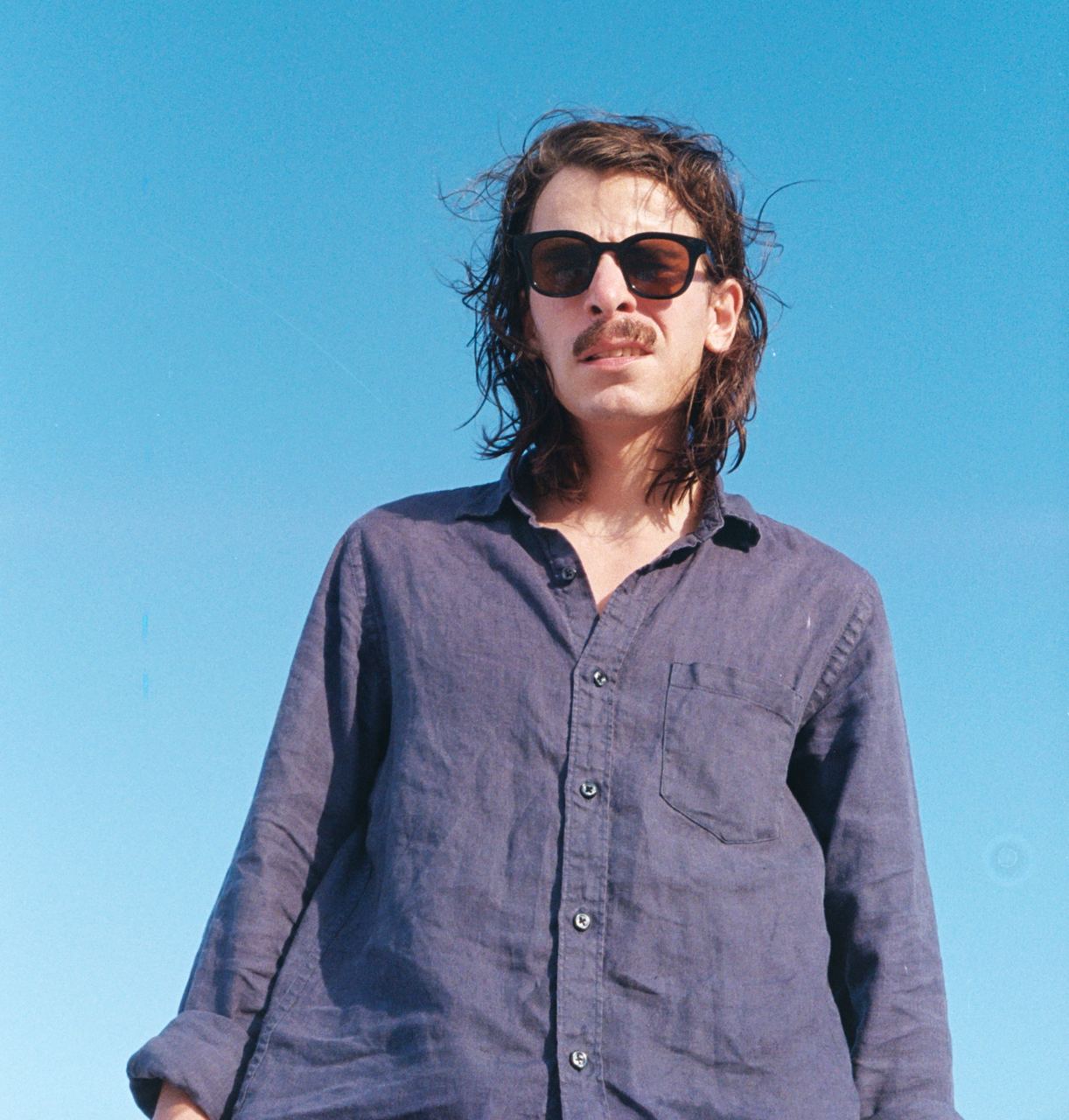 Photo of painter with a pair of brown sunglasses in a blue shirt. The background is in gradient sky blue. 