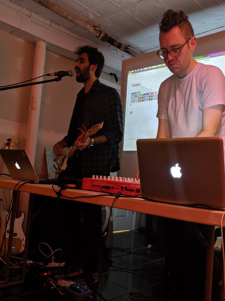 Photo of Sumanth Srinivasan and Matthew Kaney doing live coding performance with microphone, computers, electrical instrument, and MIDI controller in front of a projection screen with live code projection. 