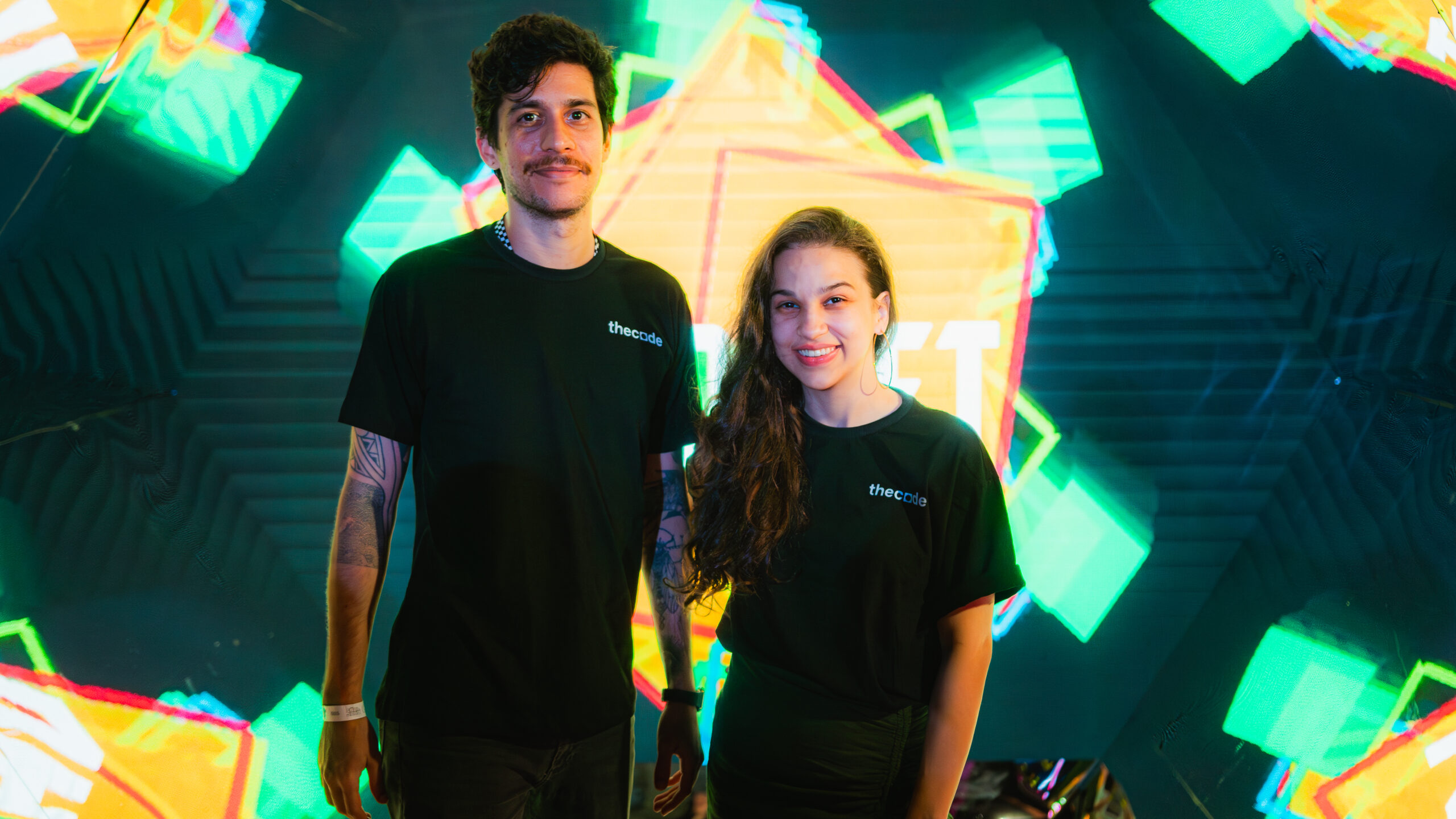 Photo of Francisco Barretto and Micaelle Lages Lucena standing together and smiling at the camera. Behind them is a big projection screen with visuals of glowing geometric shapes on a greenish background. 
