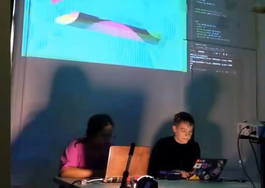Photo of this.xor.that and CougarsAreCatsToo doing live coding performance on their laptop. Behind them is a projection of live visuals and codes. 