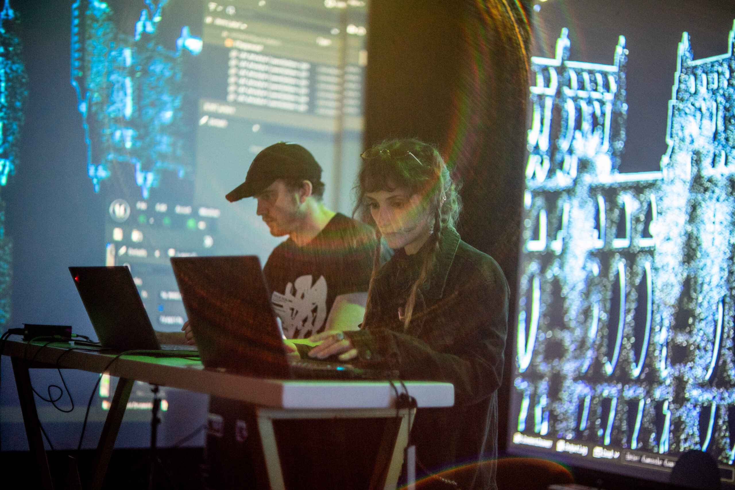 Photo of Voyde and her partner sitting behind a table and typing on their laptops, with live visuals projected on the walls behind them. 