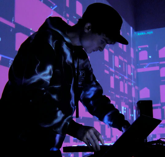 Photo of Don Hanson wearing a cap and working on a MIDI keyboard with glowing keypads in the dark. Behind him is a projection of purple background and pink geometric shapes. 