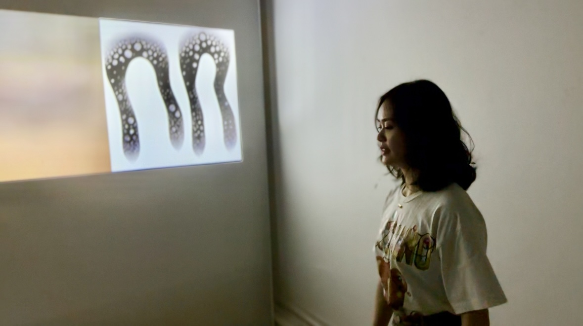 Photo of Fuguo walking in front of her projection installation inside a white room, which displays AI-generated animations. 