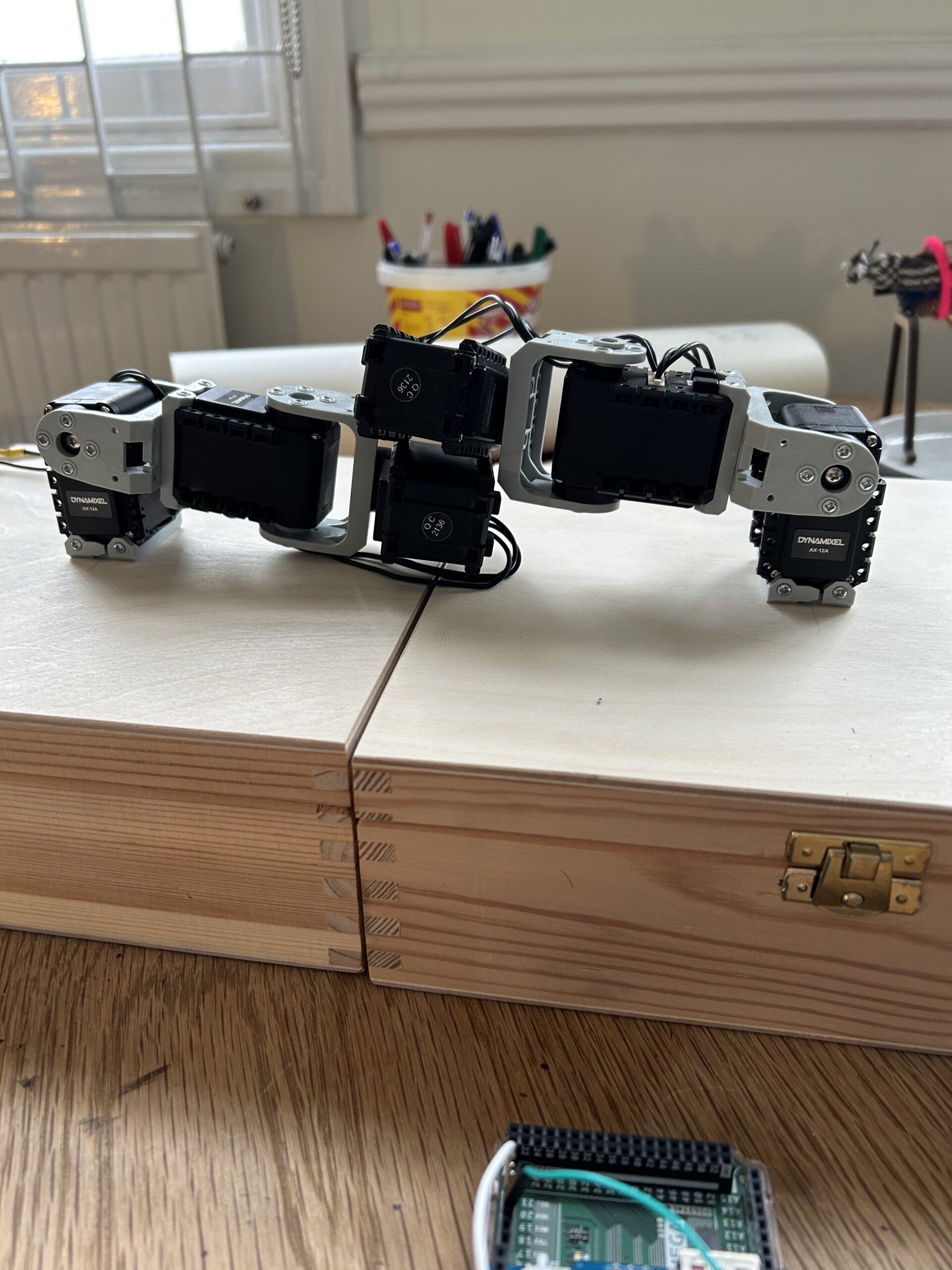 Photo of a small-scale installation consisting of two mechanical arms in black and gray, each fixed to the top of a wooden box on a wooden table. 