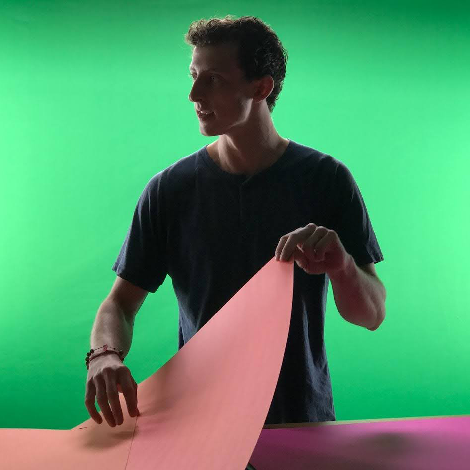 Close-up photo of Torin Blankensmith in front of a green screen, holding a piece of pink paper jam and looking to his right. 
