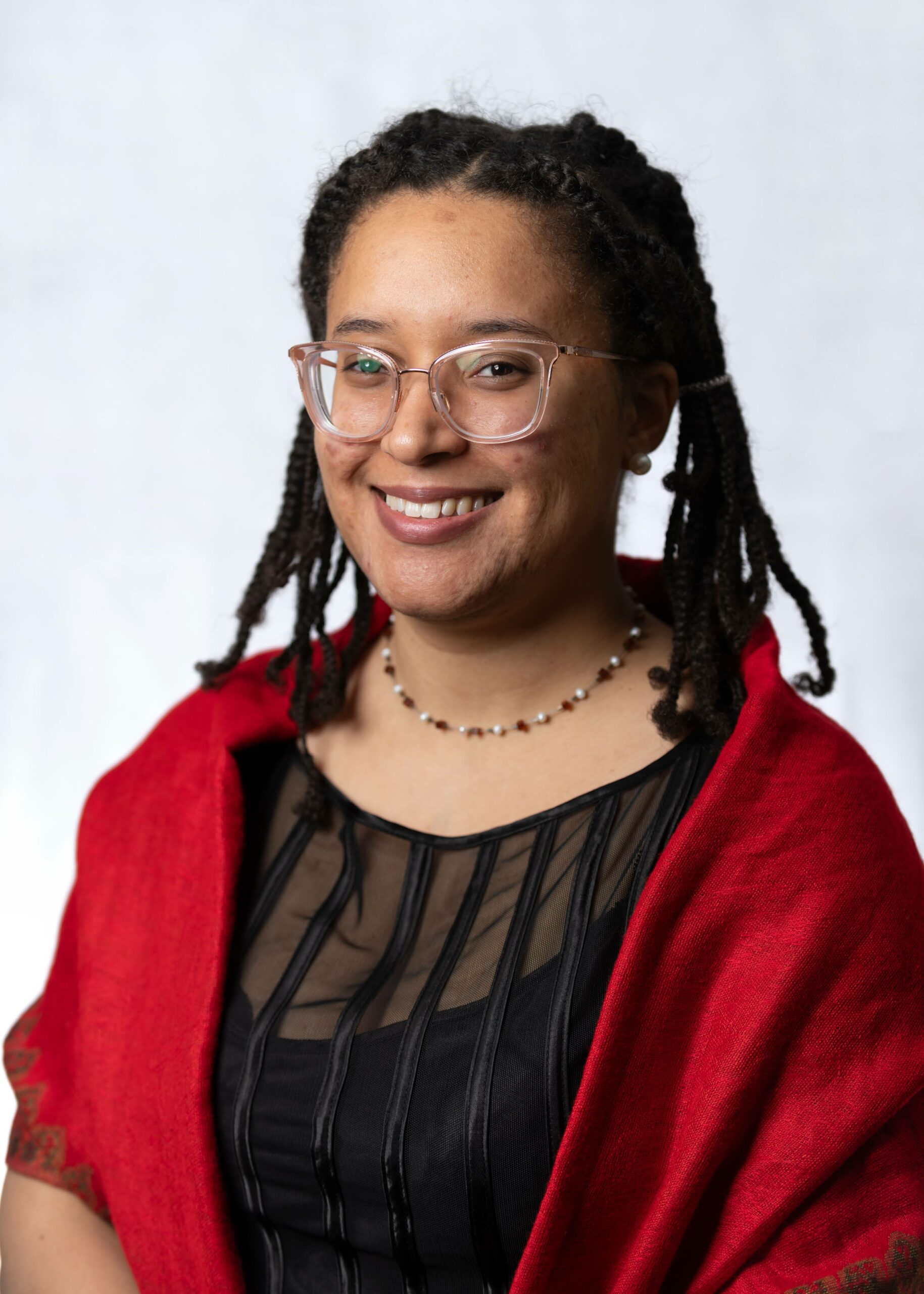 Headshot photo of CougarsAreCatsToo with a pair of glasses and in a necklace, a black shirt, and a red coat. 