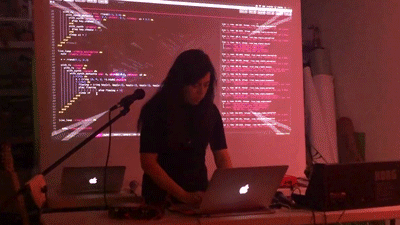GIF photo of Melody Loveless working on her laptop on a table, with her code and live visuals projected on a screen behind her. 