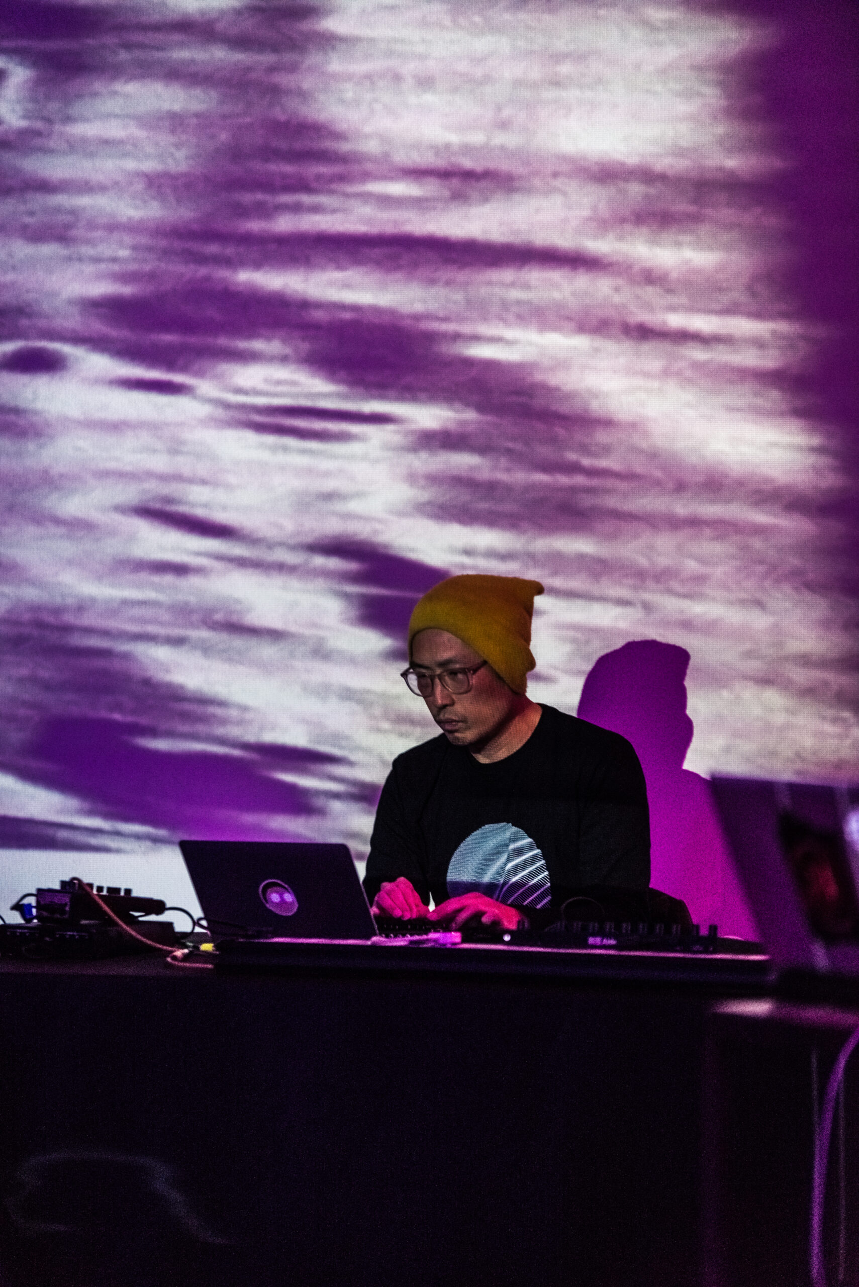 Photo of easterner typing behind a table with a laptop and mixers, and in front of a wide and tall projection screen covered in purple and white projection. 