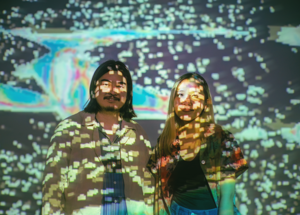 A photo of Kengchakaj & Nitcha(elekhlekha.) On the left, Kengchakaj, a Southeast Asia man with black medium-length hair and mustache.He’s wearing a blue shirt with an off-white jacket. On the right, Nitcha, a Southeast Asian woman, with medium-length bleached straight hair with black roots. She’s wearing black tank top with colorful tie-dye short sleeve shirt.  They have the particle and blue graphic from projector light shining on their face and fill the background.

Photo by Thomas J. Logan, courtesy of Institute for Electronic Arts (IEA)